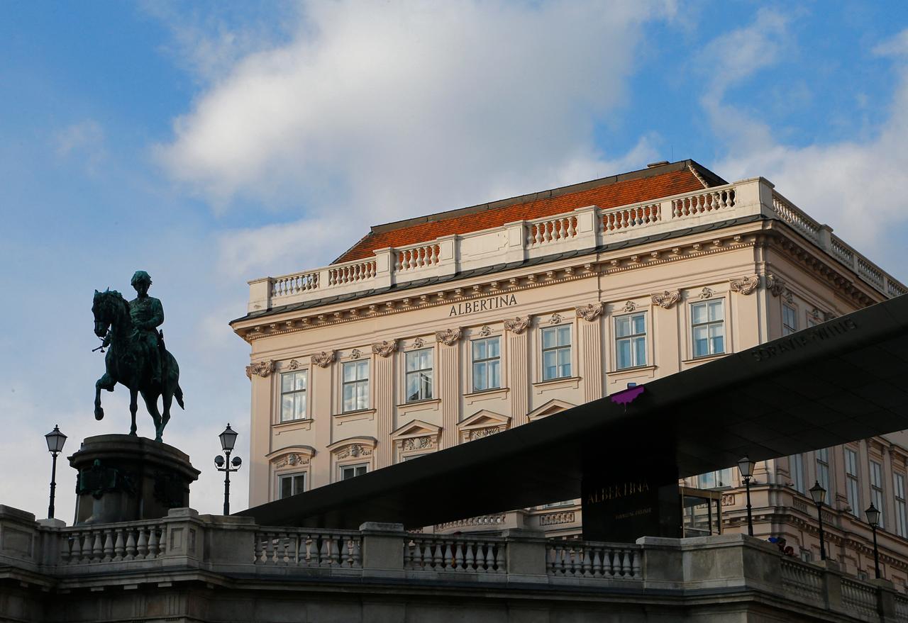 Das Albertina Museum in Wien. Das mehrstockige Museumsgebäude ist im Hintergrund. Auf einem Wandstück direkt unter dem Dach steht "ALBERTINA" geschriben. Im Vordergrund steht eine Reiterstatute als Denkmal an den Begründer der Albertina Albert Kasimir von Sachsen-Teschen.