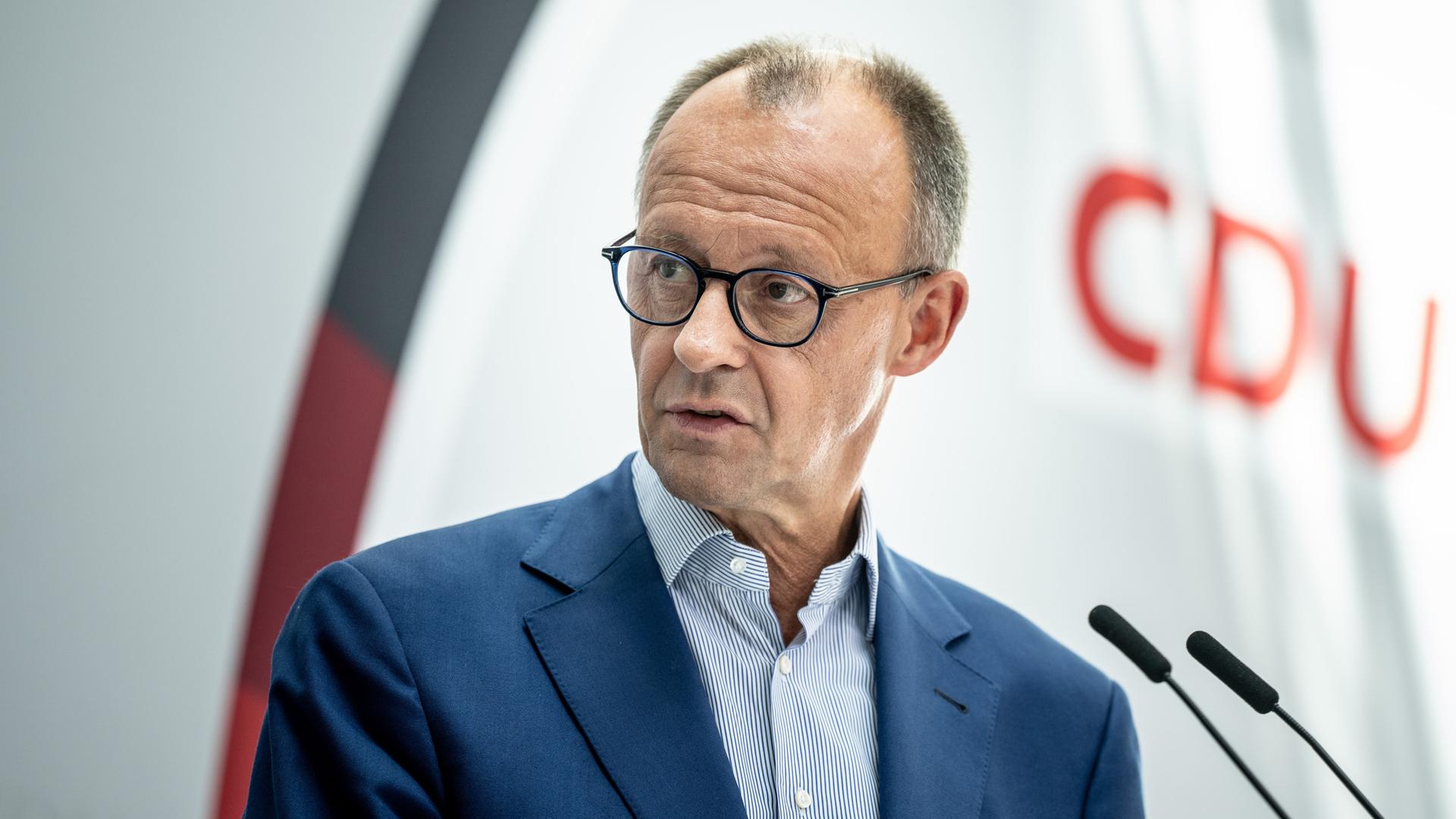 Friedrich Merz steht an einem Rednerpult, er trägt ein gemustertes Hemd offen und ohne Krawatte und ein blaues Sakko. Im Hintergrund ist rechts das Logo der CDU zu sehen.