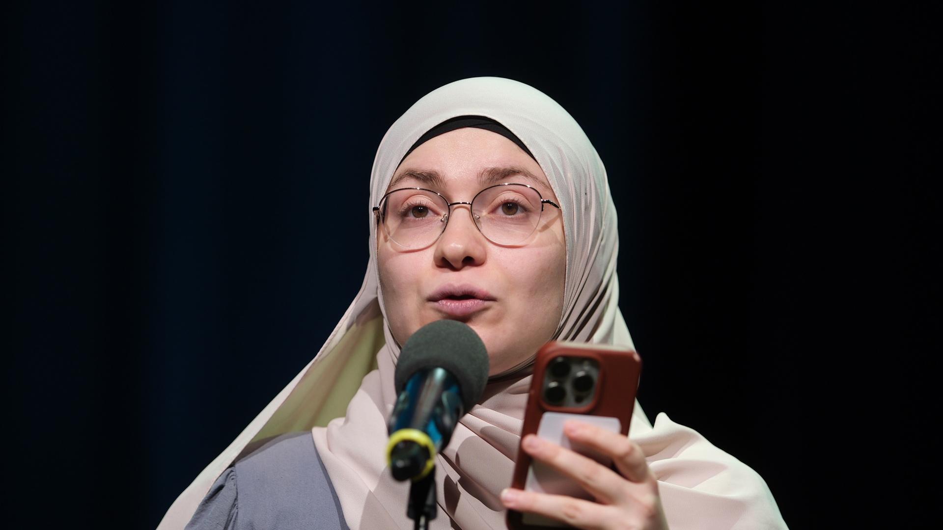 Das Bild zeigt Ayse Irem in der Stadthalle in Chemnitz beim Finale der 29. Deutschen Meisterschaften im Poetry-Slam. Sie steht auf der Bühne hinter einem Mikrofon und hält während ihrer Präsentation ihr Handy in der Hand. Der Hintergrund ist dunkel.