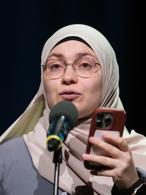 Das Bild zeigt Ayse Irem in der Stadthalle in Chemnitz beim Finale der 29. Deutschen Meisterschaften im Poetry-Slam. Sie steht auf der Bühne hinter einem Mikrofon und hält während ihrer Präsentation ihr Handy in der Hand. Der Hintergrund ist dunkel.