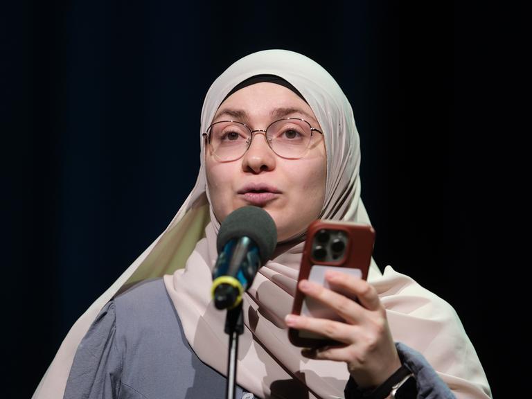 Das Bild zeigt Ayse Irem in der Stadthalle in Chemnitz beim Finale der 29. Deutschen Meisterschaften im Poetry-Slam. Sie steht auf der Bühne hinter einem Mikrofon und hält während ihrer Präsentation ihr Handy in der Hand. Der Hintergrund ist dunkel.