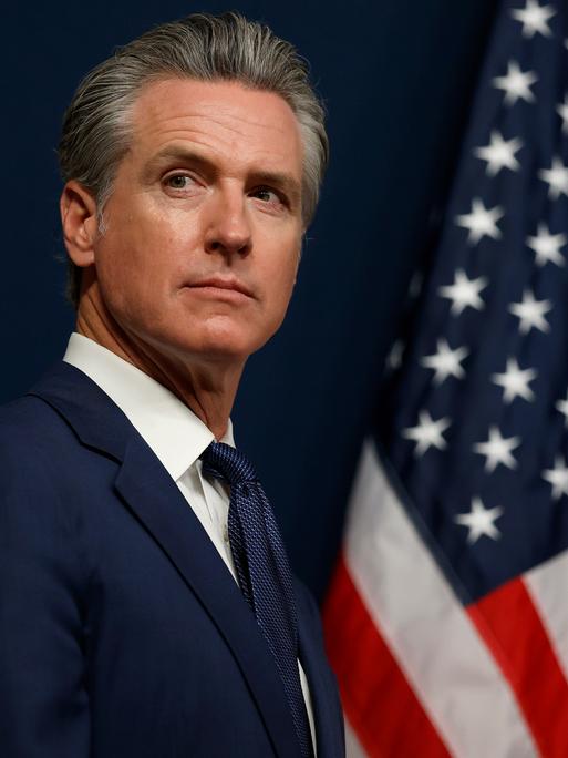 Kaliforniens Gouverneur Gavin Newsom trägt einen blauen Anzug und steht zwischen zwei amerikanischen Flaggen.