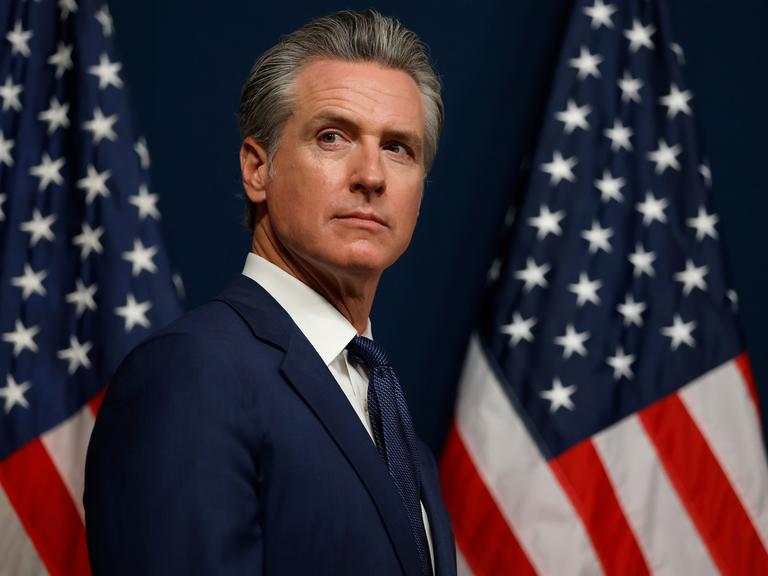 Kaliforniens Gouverneur Gavin Newsom trägt einen blauen Anzug und steht zwischen zwei amerikanischen Flaggen.