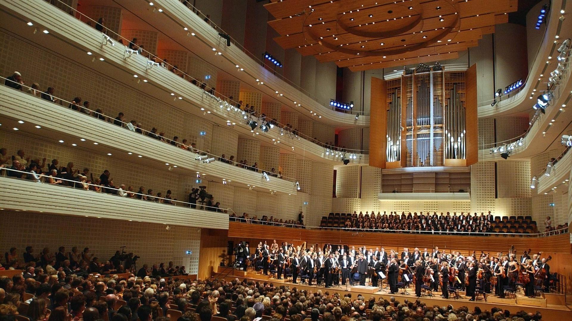 Ein voll besetzter Konzertsaal mit großer Orgel und beleuchteten Rängen. Das Orchester und der Chor stehen. Ein voll besetzter Konzertsaal mit großer Orgel und beleuchteten Rängen. Das Orchester und der Chor stehen.
