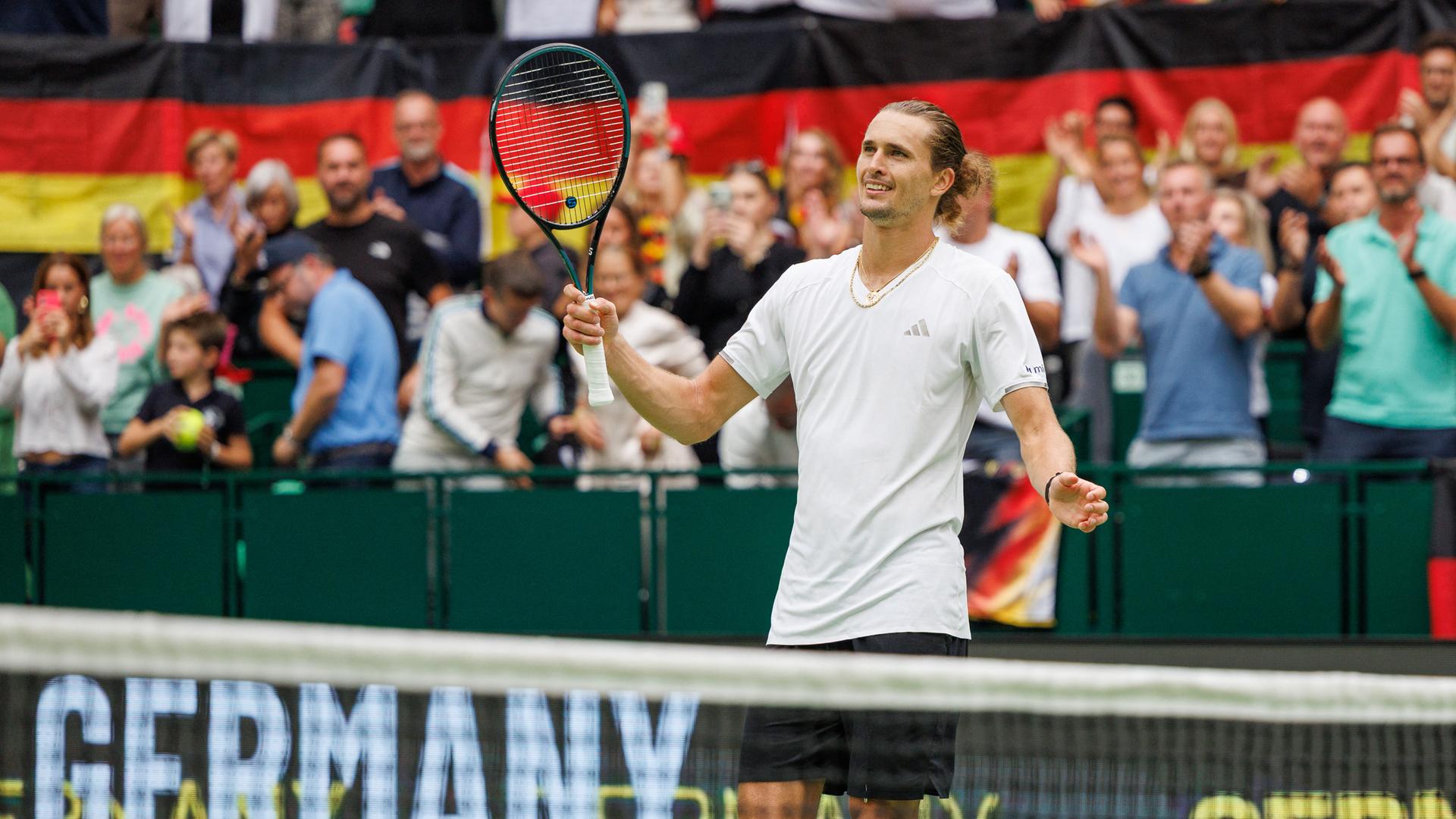 Alexander Zverev steht hinter einem Tennisnetz, hebt den Schläger und lächelt. 