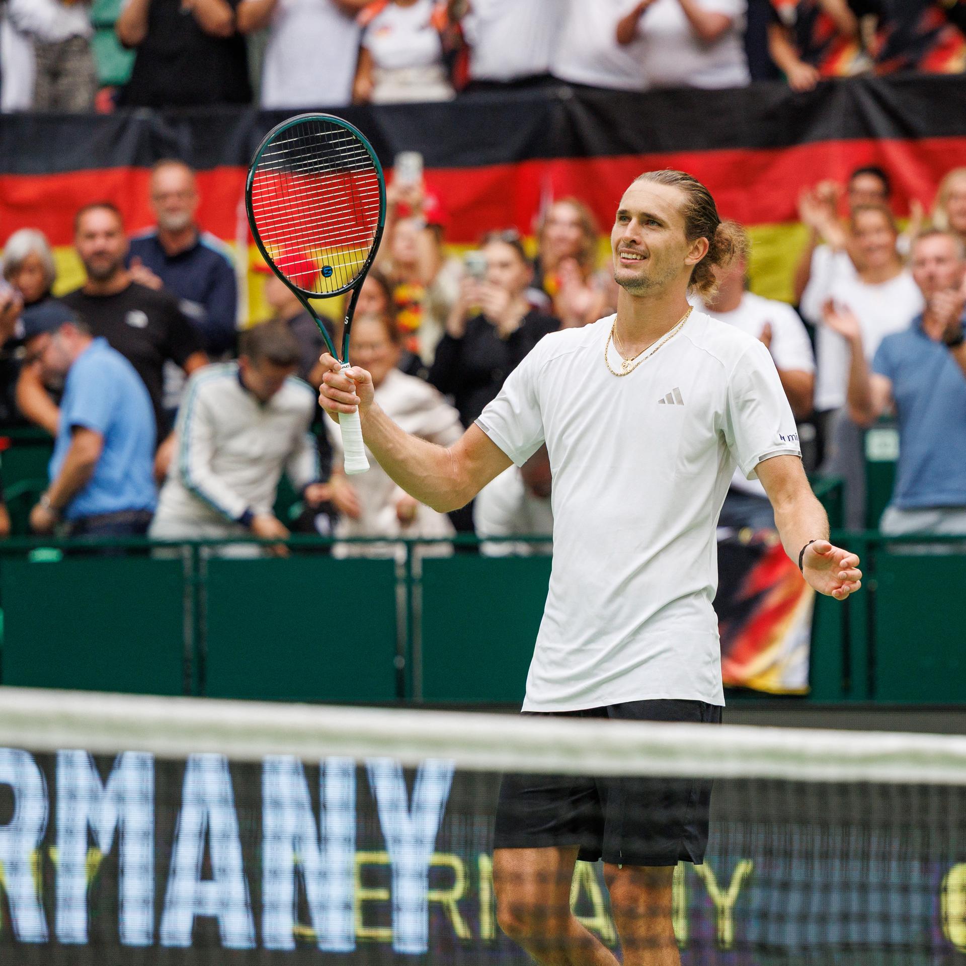 Alexander Zverev steht hinter einem Tennisnetz, hebt den Schläger und lächelt. 