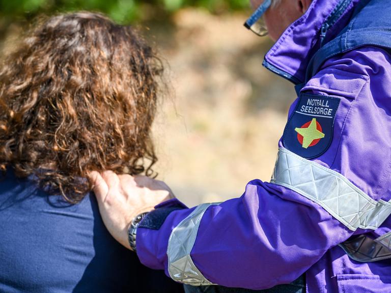 Ein Notfallseelsorger kniet neben einer Frau und legt seine Hand auf ihre Schulter. Auf der violetten Jacke ist das gelbe Kreuz der Notfallseelsorge zu erkennen.