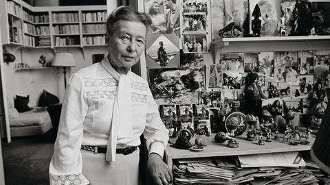 Simone de Beauvoir steht in ihrer Wohnung voller Bücher und schaut in die Kamera