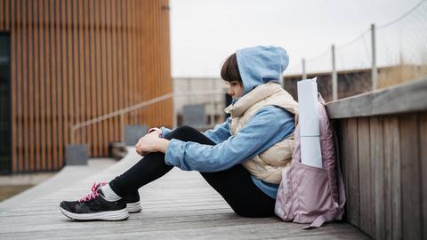 Ein Mädchen hockt mit ins Gesicht gezogener Kapuze und angezogenen Knien auf einer breiten Holztreppe. Neben ihr steht ein rosafarbener Rucksack.