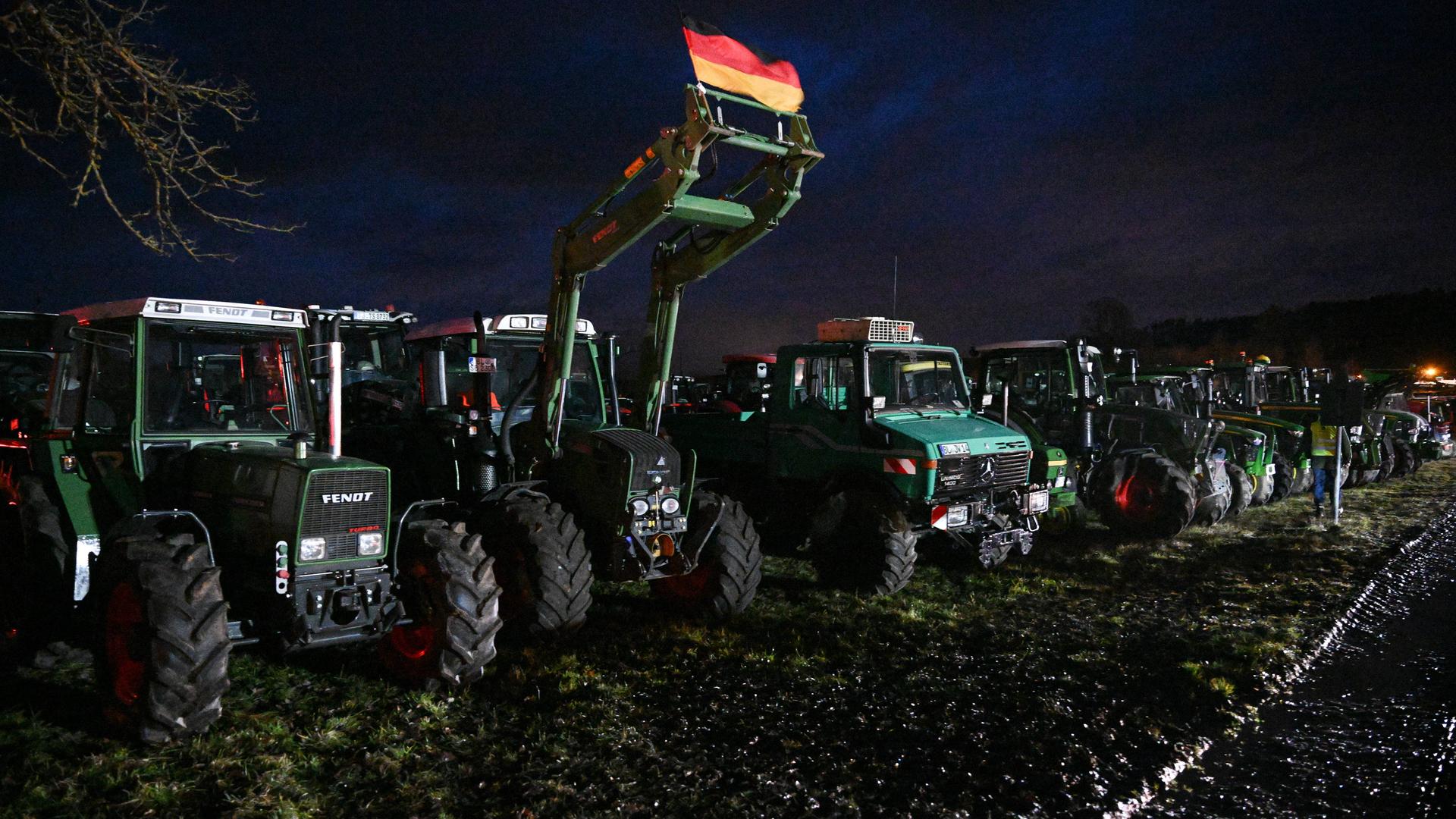 Viele Traktoren und ein Unimog stehen auf einem Feld. An einem ist eine Deutschlandfahne angebracht.