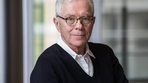 László Földényi sitzt vor einer großen Glasscheibe, trägt eine Hornbrille und schaut freundlich in die Kamera