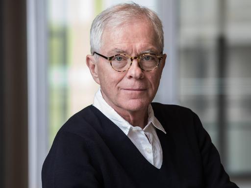 László Földényi sitzt vor einer großen Glasscheibe, trägt eine Hornbrille und schaut freundlich in die Kamera
