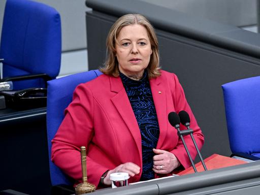 Bärbel Bas, Präsidentin des Deutschen Bundestages, trägt ein dunkelrosafarbenes Jackett. Sie sitzt auf einem Stuhl hinter zwei Mikrofonen, vor ihr steht eine umgedrehte Glocke.
