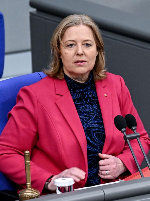 Bärbel Bas, Präsidentin des Deutschen Bundestages, trägt ein dunkelrosafarbenes Jackett. Sie sitzt auf einem Stuhl hinter zwei Mikrofonen, vor ihr steht eine umgedrehte Glocke.