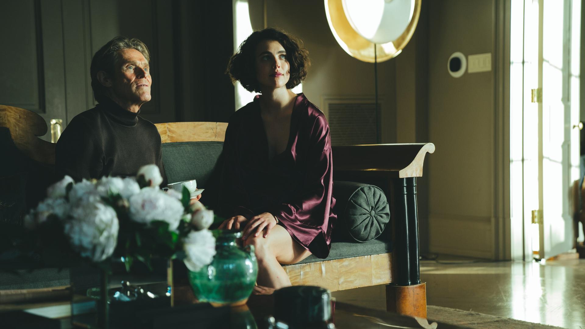 Willem Dafoe and Margaret Qualley sitzen in einer Szene des Films "Kinds Of Kindness" von Yorgos Lanthimos nebeneinander auf einem Sofa.