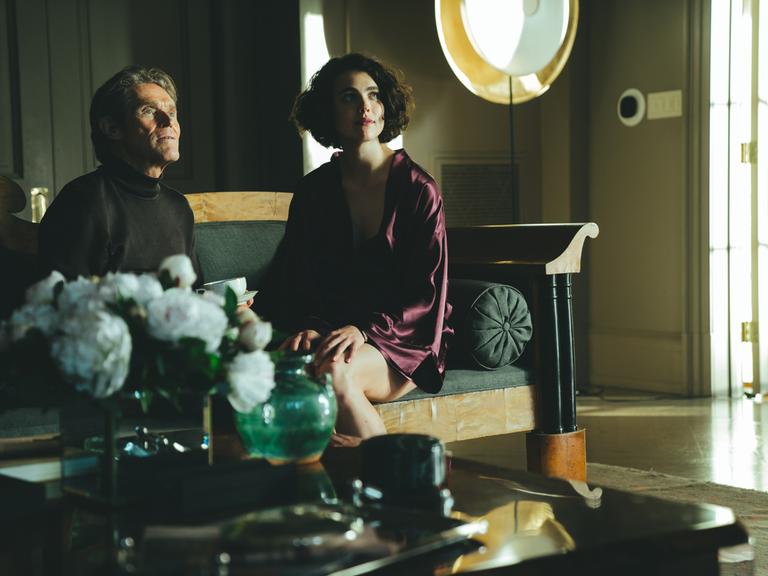 Willem Dafoe and Margaret Qualley sitzen in einer Szene des Films "Kinds Of Kindness" von Yorgos Lanthimos nebeneinander auf einem Sofa.