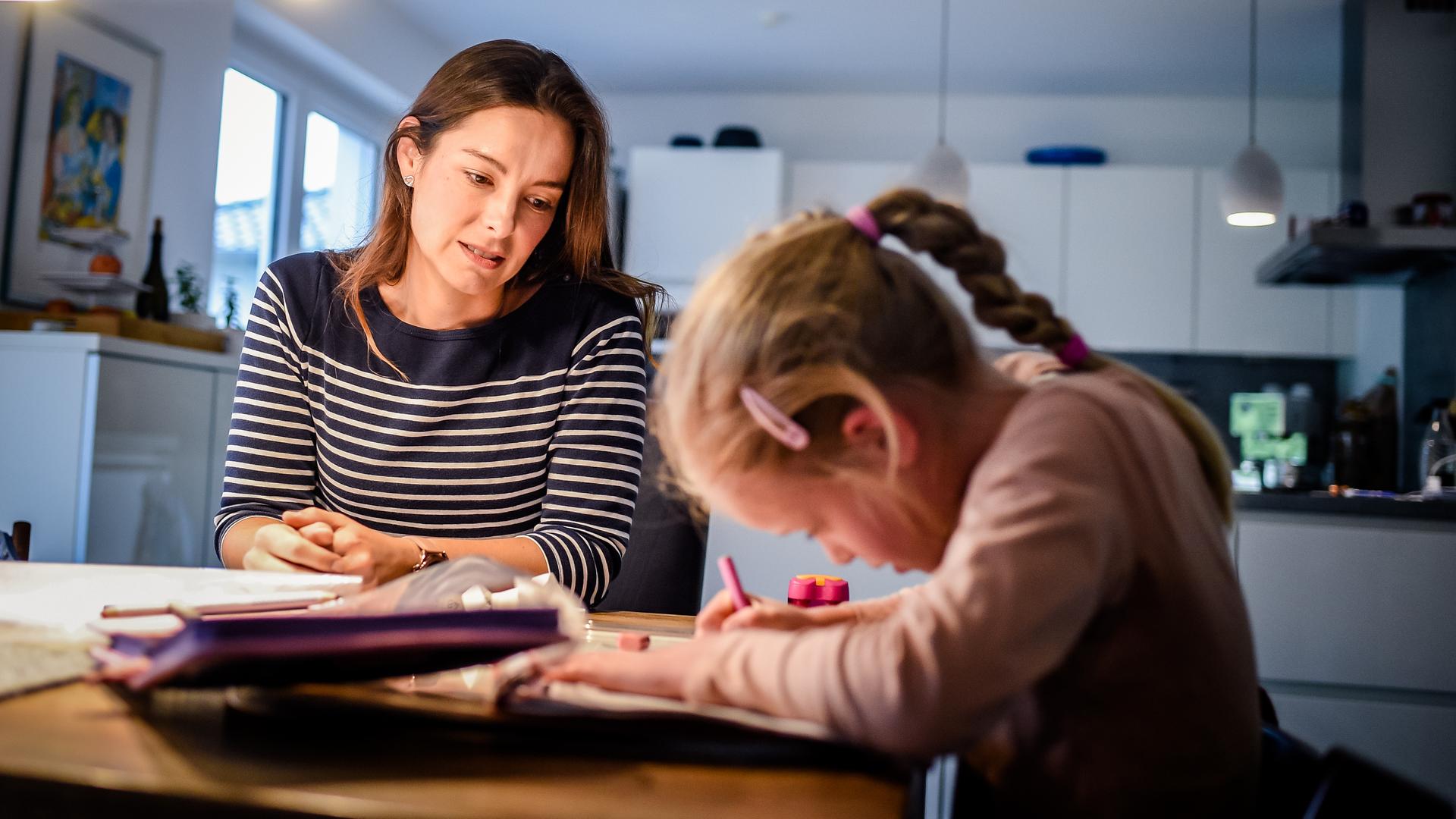 Eine Frau sitzt mit ihrer Tochter am Küchentisch. Das Kind macht Hausaufgaben.