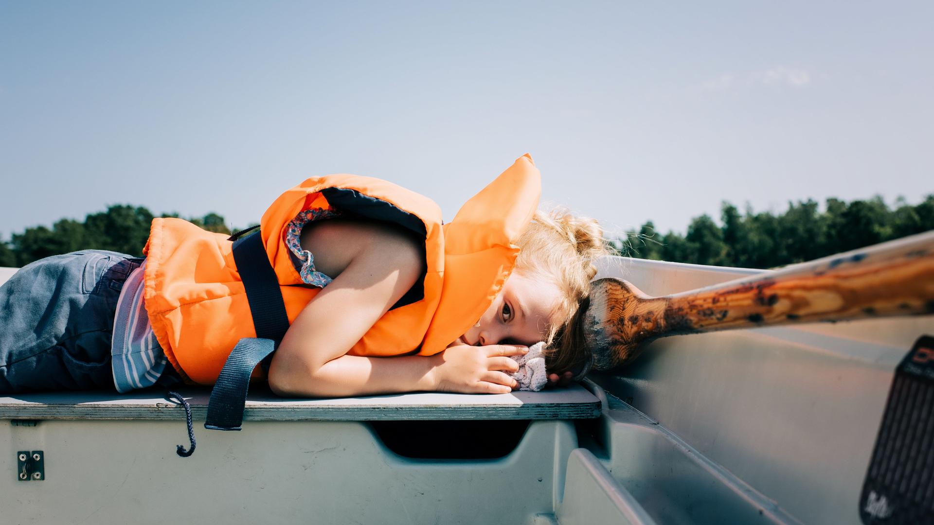 Ein Kind liegt in einem Ruderboot. Es trägt eine neonorangefarbene Rettungsweste.