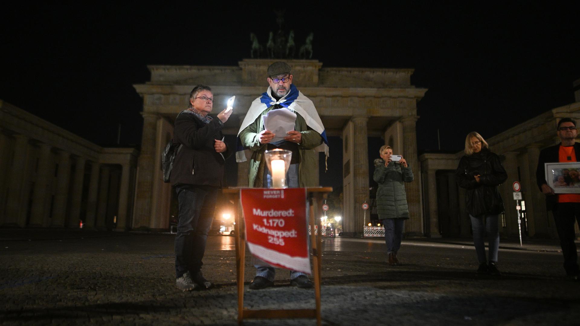 Ein Mann steht vor dem Brandenburger Tor in Berlin. Es ist dunkel. Eine Frau macht ihm Licht mit ihrem Handy. Der Mann liest etwas vor. Um seine Schultern hängt die Flagge von Israel. Ein Mann steht vor dem Brandenburger Tor in Berlin. Es ist dunkel. Eine Frau macht ihm Licht mit ihrem Handy. Der Mann liest etwas vor. Um seine Schultern hängt die Flagge von Israel.