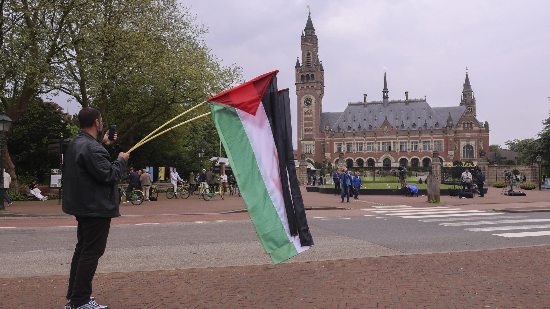 Ein pro-palästinensischer Demonstrant steht vor dem IGH in Den Haag und hält zwei Palästina-Flaggen hoch.