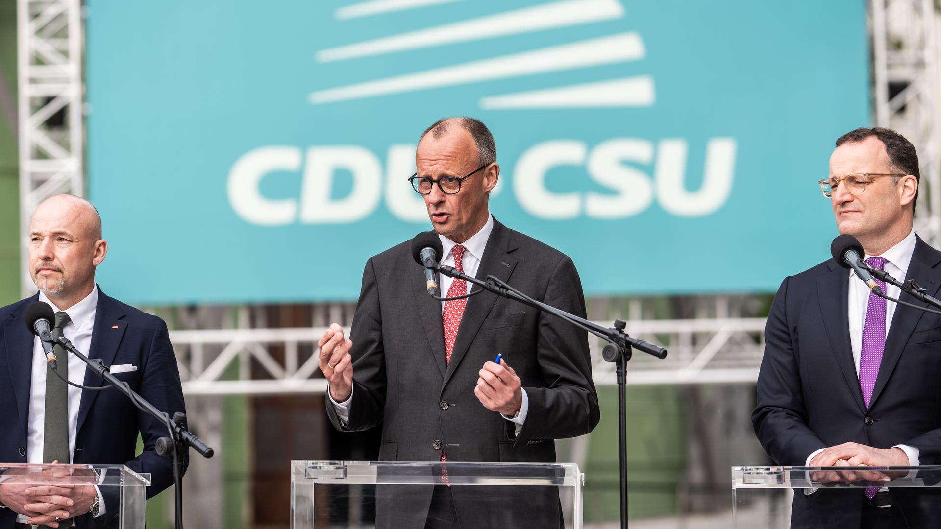 Bundeskanzler Friedrich Merz (M, CDU) steht zwischen Jens Spahn und Alexander Hoffmann und spricht in ein Mikrofon. Dahinter eine lindgrüne Wand mit den Logos beider Parteien.