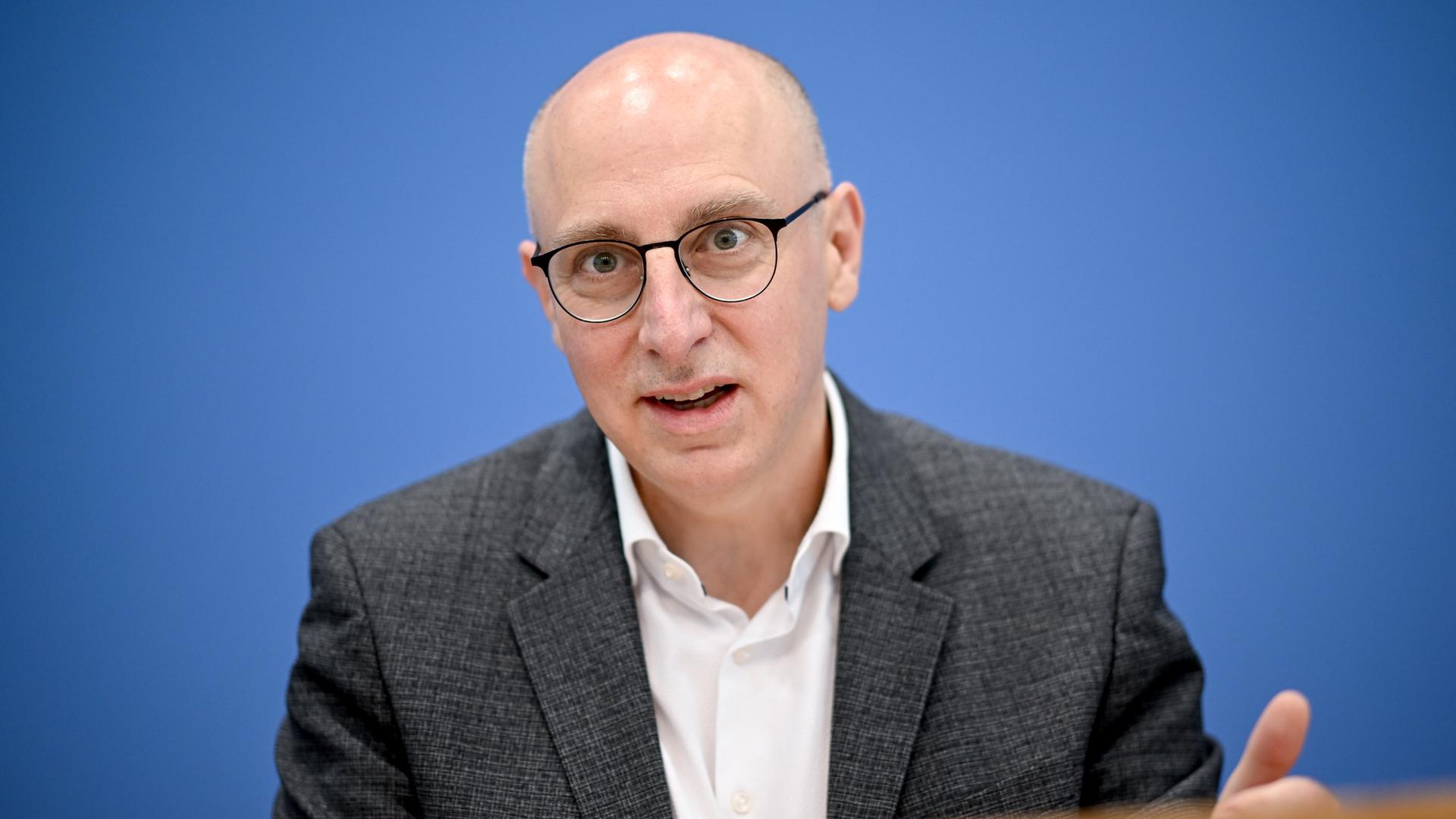 Sinan Selen sitzt vor einer blauen Wand und gestikuliert.