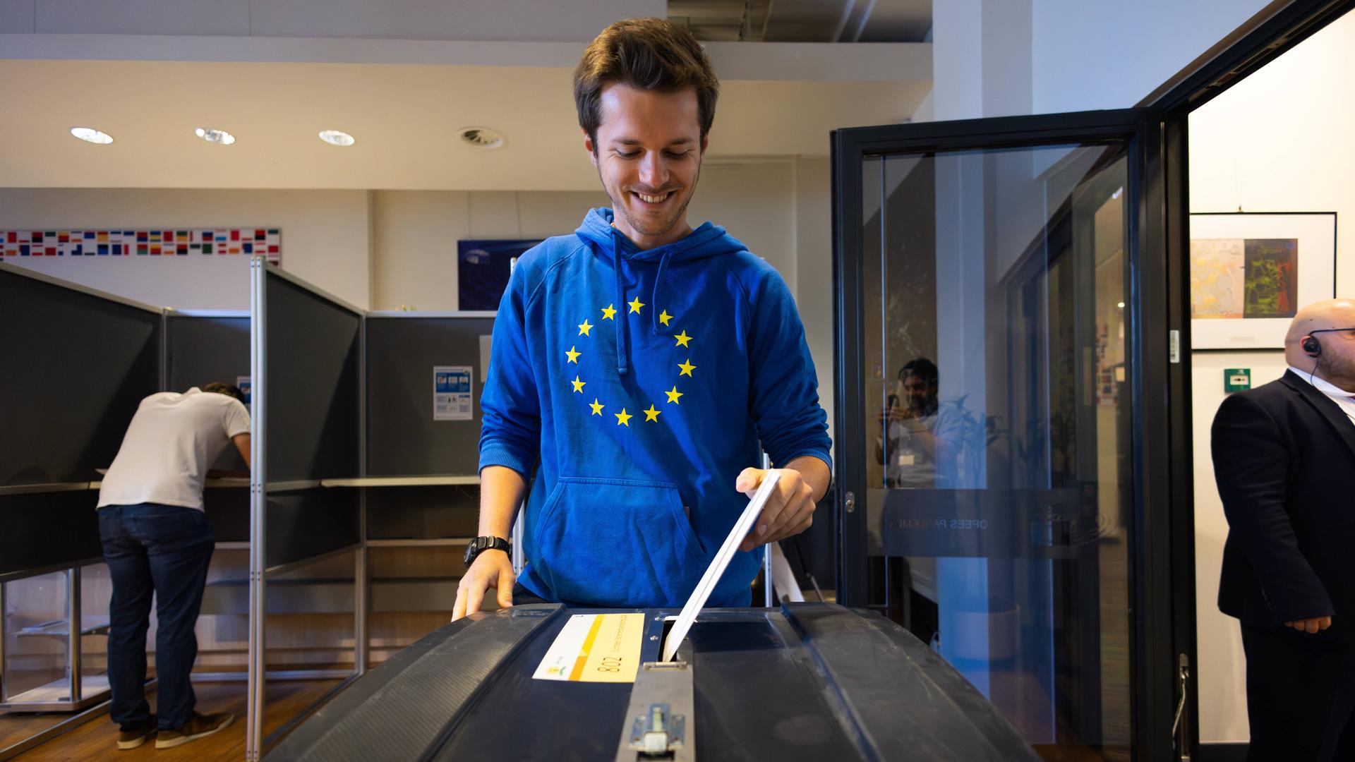 Den Haag, Niederlande: Ein Mann in blauem Pulli mit den gelben Sternen der EU steckt seinen Wahlzettel in die Wahlurne. Im Hintergrund steht ein anderer Mann in einer Wahlkabine.