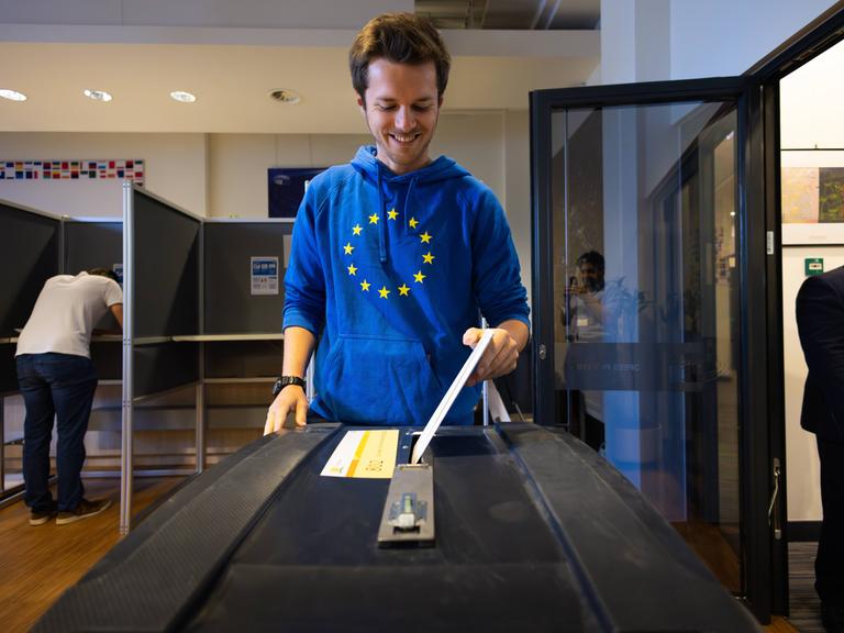 Den Haag, Niederlande: Ein Mann in blauem Pulli mit den gelben Sternen der EU steckt seinen Wahlzettel in die Wahlurne. Im Hintergrund steht ein anderer Mann in einer Wahlkabine.