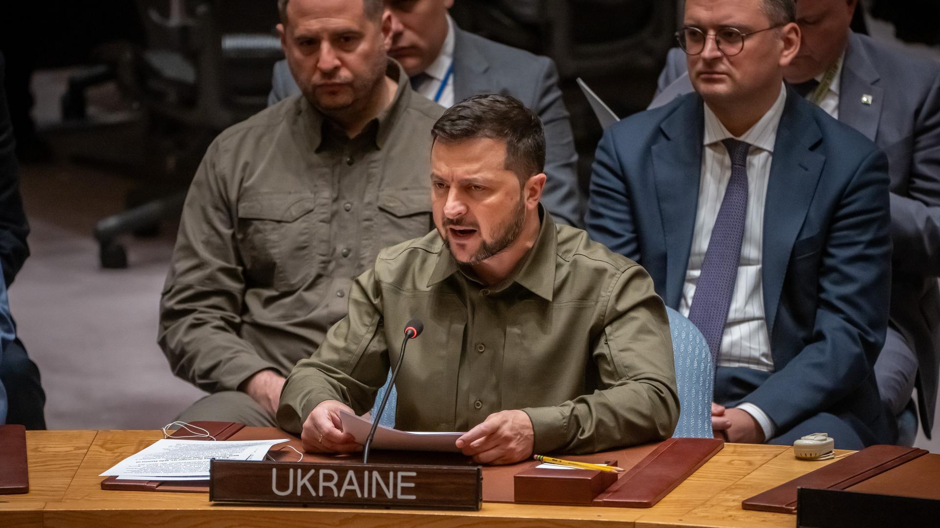 Selenskyj hält seine Ansprache im Sitzen. Er sitzt hinter einem Mikrofon und einem Schild mit den Worten UKRAINE.