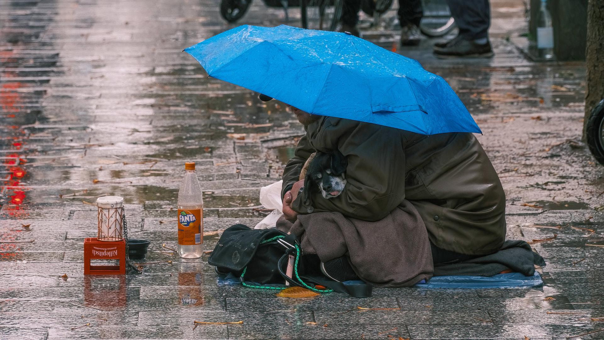 Ein Obdachloser sitzt mit seinem kleinen Hund auf einem verregneten Gehweg. Ein Regenschirm bietet den beiden notdürftigen Schutz.