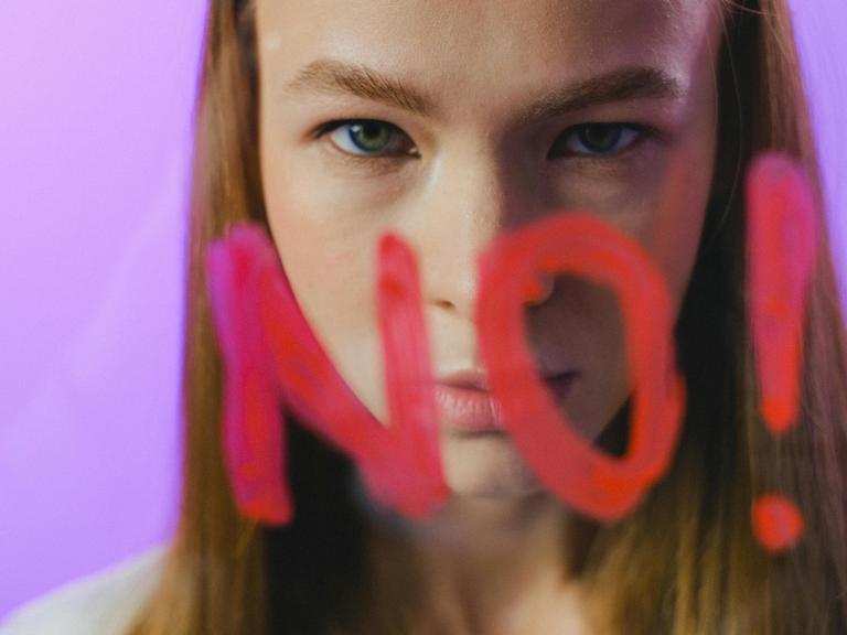 Eine junge Frau mit grünen Augen und rotbraunen Haaren fixiert die Kamera über dem Schriftzug "No!", der in großen roten Buchstaben auf einer Scheibe notiert wurde.