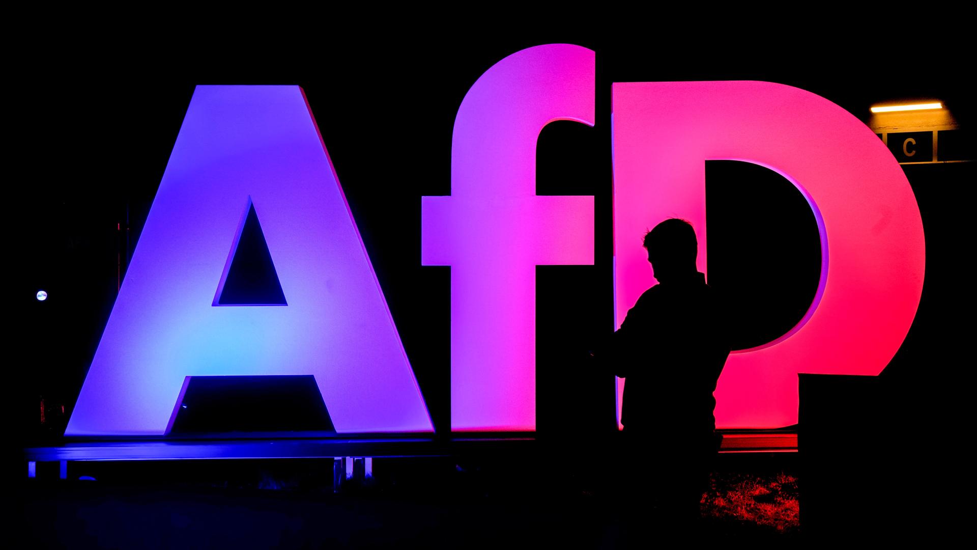 Die Silhouette einer Person vor einem rosa-violett beleuchteten AfD-Logo in einem dunklen Raum.
