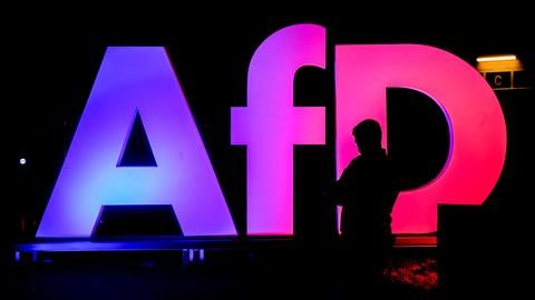 Die Silhouette einer Person vor einem rosa-violett beleuchteten AfD-Logo in einem dunklen Raum.