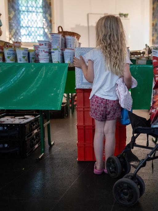 Ein Mädchen schaut sich während der Ausgabezeit in einer Ausgabestelle der Berliner Tafel die Auslage von Lebensmitteln an. SIe ist von hinten zu sehen und neben ihr steht ein Kinderwagen.
