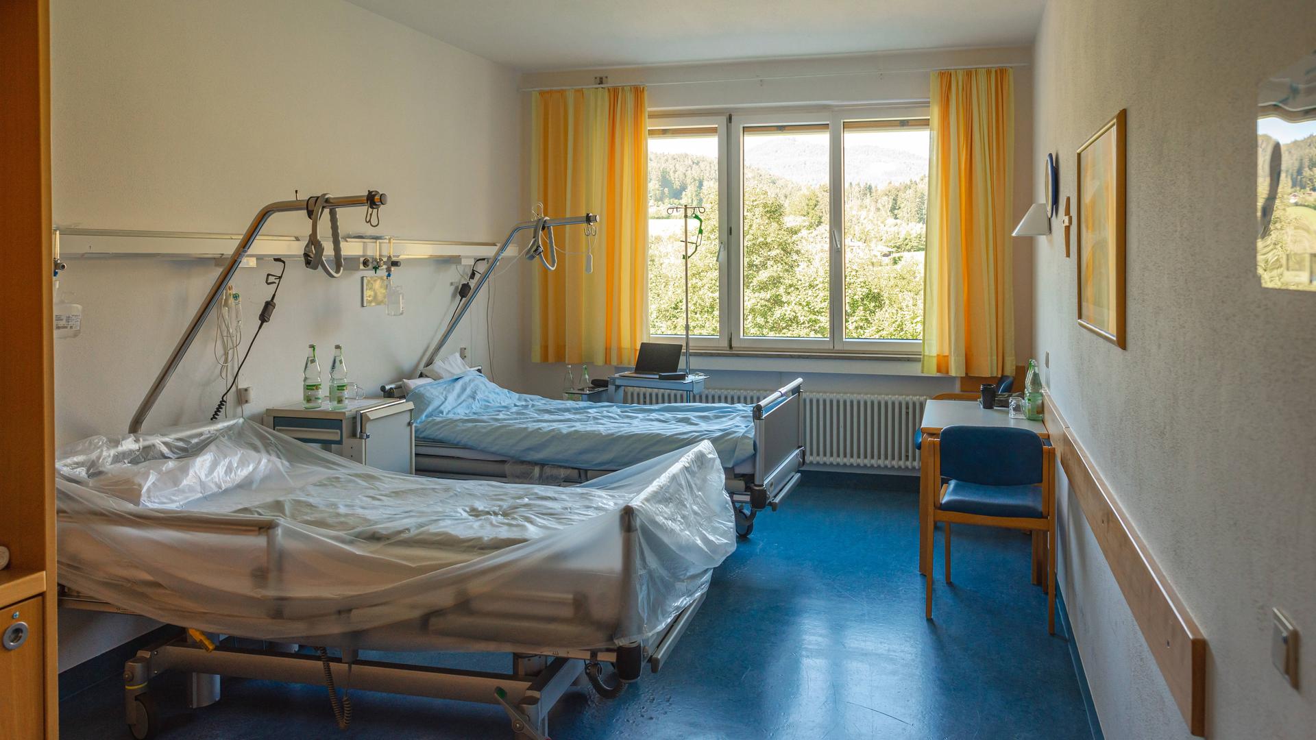 Der Blick in ein Krankenhauszimmer zeigt zwei leere Betten und ein Fenster mit Ausblick ins Grüne. Der Blick in ein Krankenhauszimmer zeigt zwei leere Betten und ein Fenster mit Ausblick ins Grüne.
