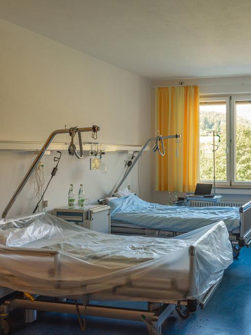 Der Blick in ein Krankenhauszimmer zeigt zwei leere Betten und ein Fenster mit Ausblick ins Grüne.