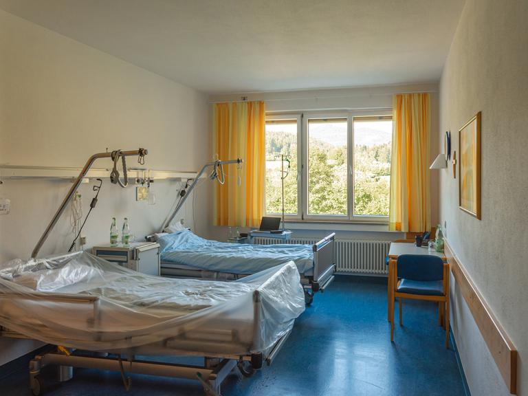 Der Blick in ein Krankenhauszimmer zeigt zwei leere Betten und ein Fenster mit Ausblick ins Grüne.