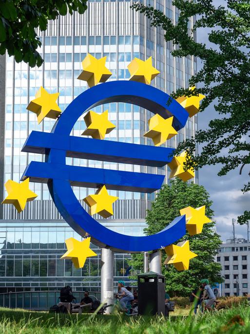 Vor einem Bürohaus steht eine Skulptur in Form eines Euro-Zeichens. Um dieses herum sind gelbe Sterne. Es steht vor der  Zentrale der Europäischen Zentralbank in Frankfurt am Main. 