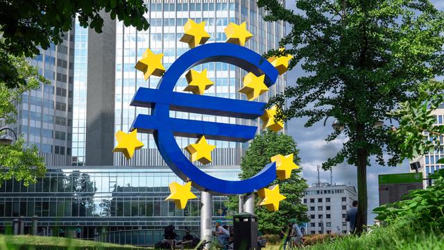 Vor einem Bürohaus steht eine Skulptur in Form eines Euro-Zeichens. Um dieses herum sind gelbe Sterne. Es steht vor der  Zentrale der Europäischen Zentralbank in Frankfurt am Main. 