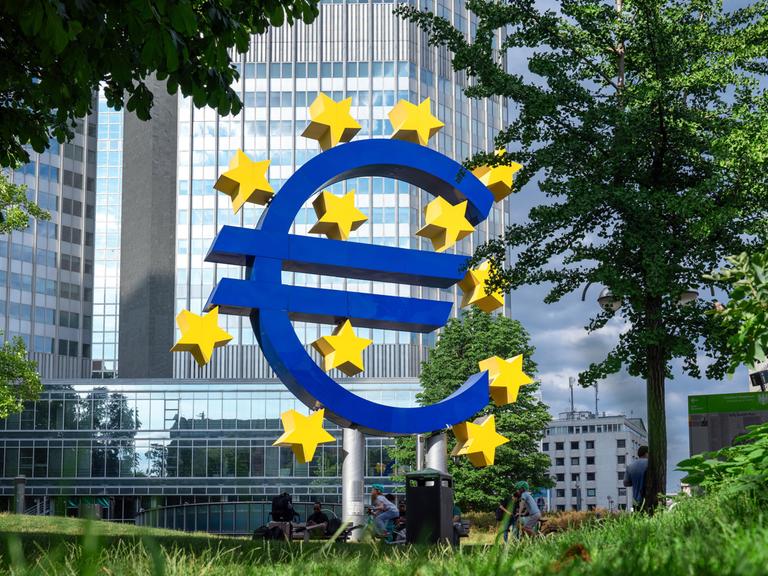 Vor einem Bürohaus steht eine Skulptur in Form eines Euro-Zeichens. Um dieses herum sind gelbe Sterne. Es steht vor der  Zentrale der Europäischen Zentralbank in Frankfurt am Main. 