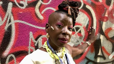 Eine Frau steht vor einer bunt besprayten Wand. Sie lehnt mit ihrer linken Hand gegen die Wand. Ihre Haare hat sie kunstvoll in die Höhe frisiert. Sie trägt auffälliges Make-Up und Schmuck.