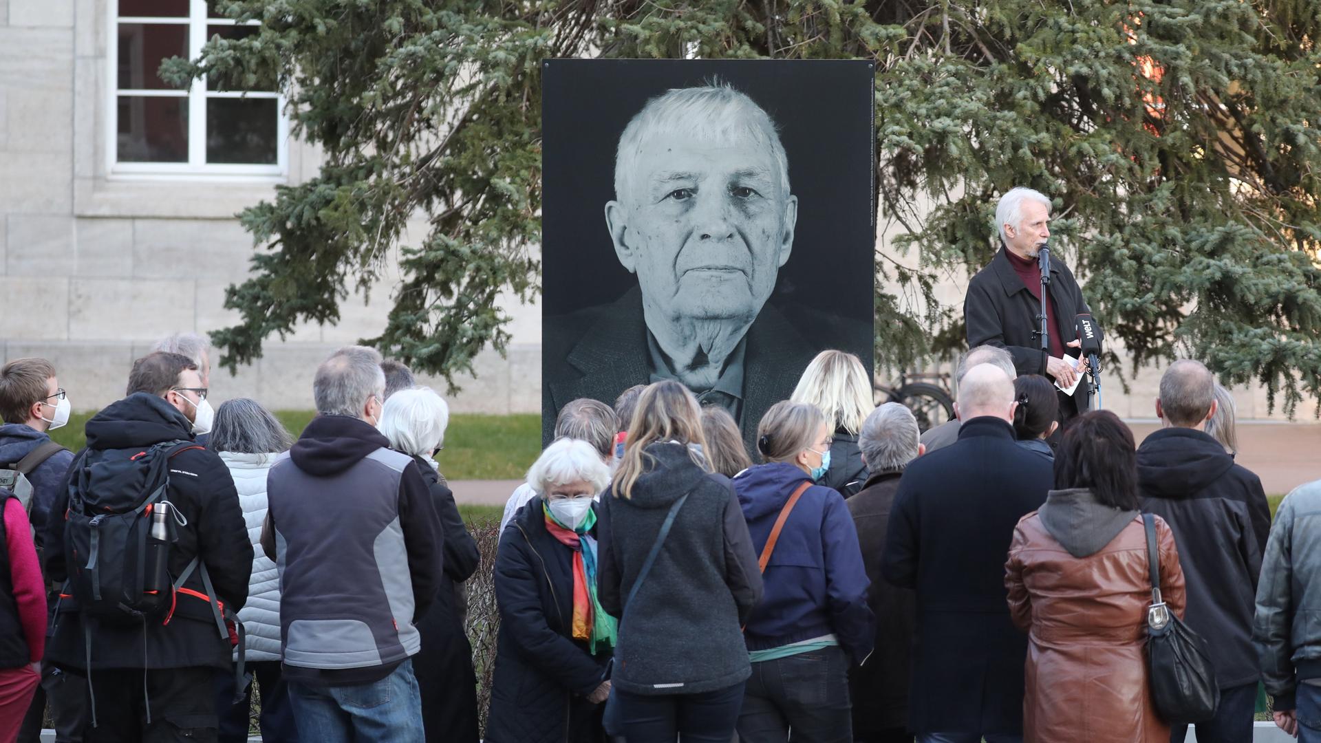 Ein schwarz-weiß Portrait von Boris Romantschenko ist vor einem Baum aufgestellt. Eine Menschenmenge steht davor. 