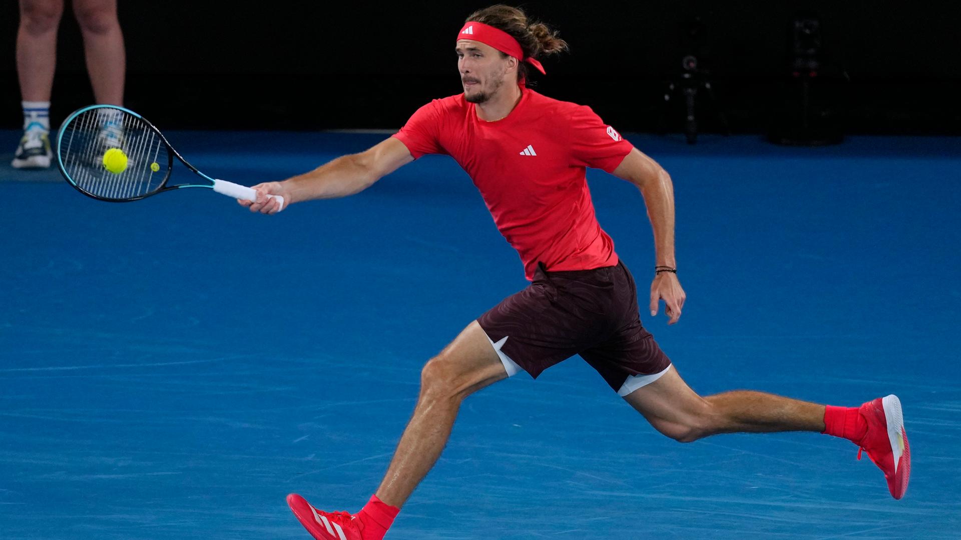 Zverev im roten Shirt und mit schwarzer Hose spielt im Laufen einen Ball.