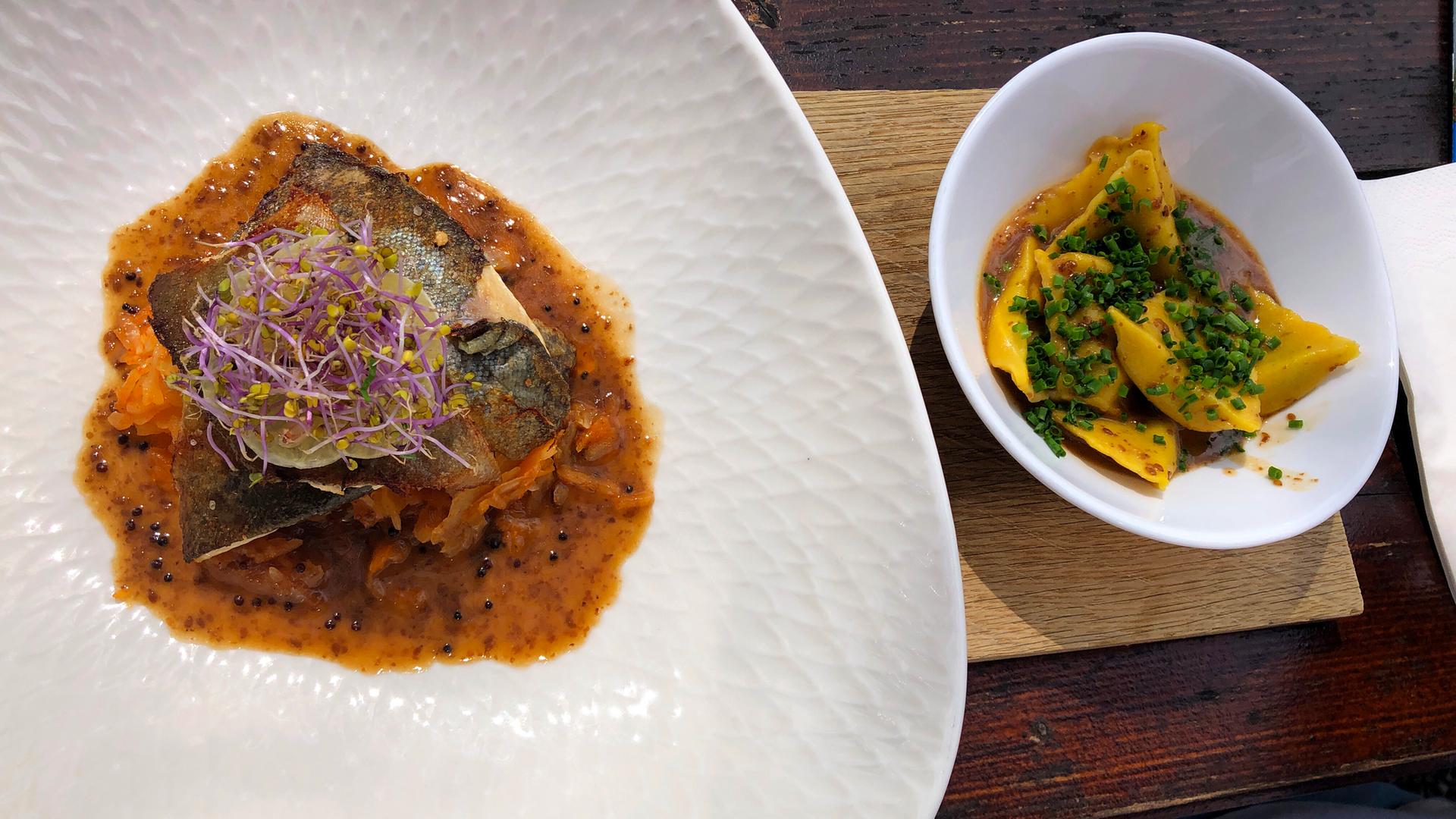 Gebratene Forellenfilets und bayrisches Kim Chi mit Teigtaschen und Sesam sind in zwei weißen Tellern angerichtet