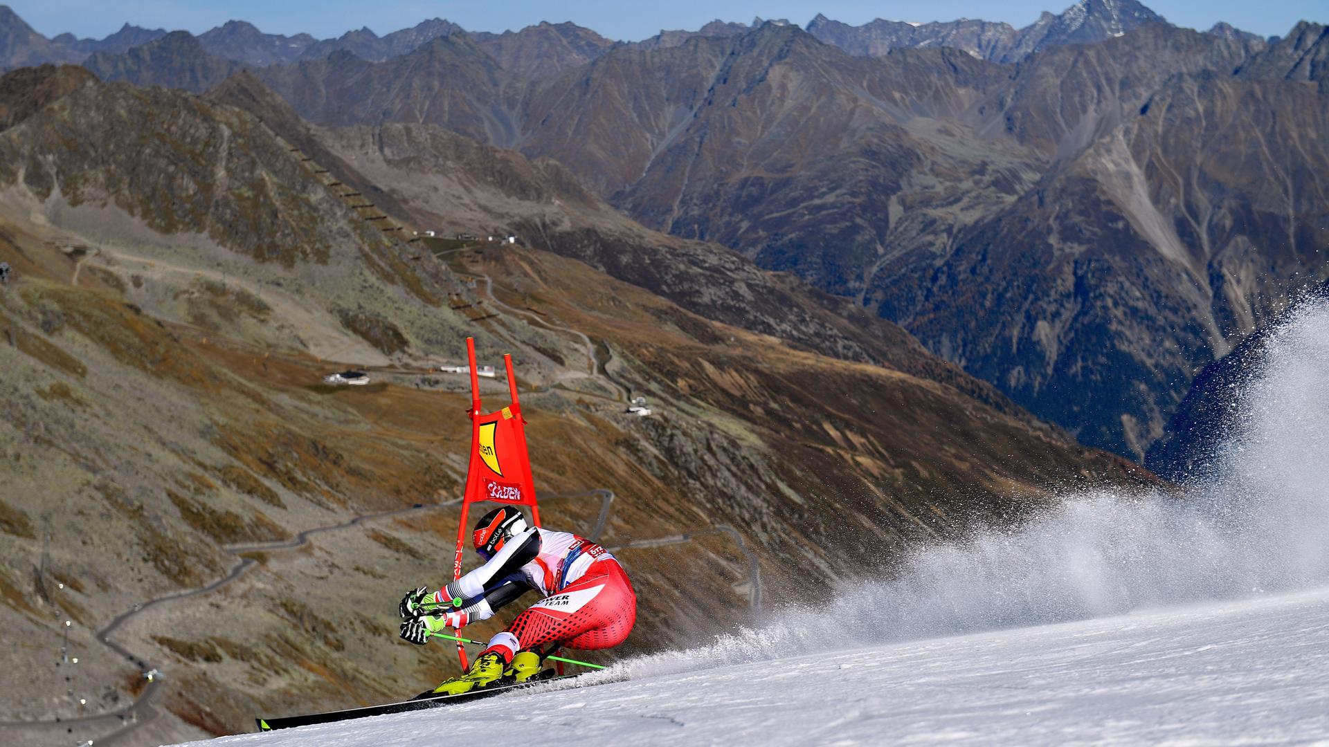 Ein Skifahrer fährt Slalom auf einer künstlich angelegten Piste. Im Hintergrund sind man grau-braune Alpenhänge. 