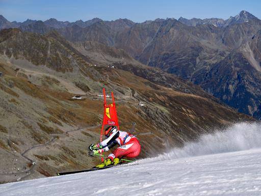 Ein Skifahrer fährt Slalom auf einer künstlich angelegten Piste. Im Hintergrund sind man grau-braune Alpenhänge. 