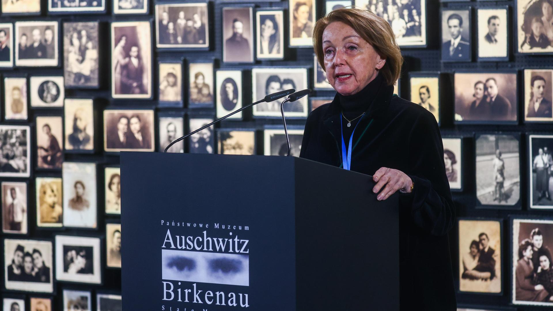 Eva Umlauf steht vor einer Wand mit Schwarz-weiß-Porträtfotos an einem Rednerpult mit der Aufschrift "Auschwitz Birkenau".