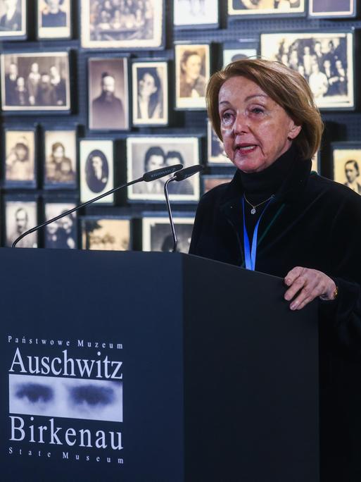 Eva Umlauf steht vor einer Wand mit Schwarz-weiß-Porträtfotos an einem Rednerpult mit der Aufschrift "Auschwitz Birkenau".