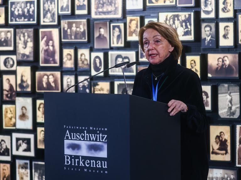 Eva Umlauf steht vor einer Wand mit Schwarz-weiß-Porträtfotos an einem Rednerpult mit der Aufschrift "Auschwitz Birkenau".