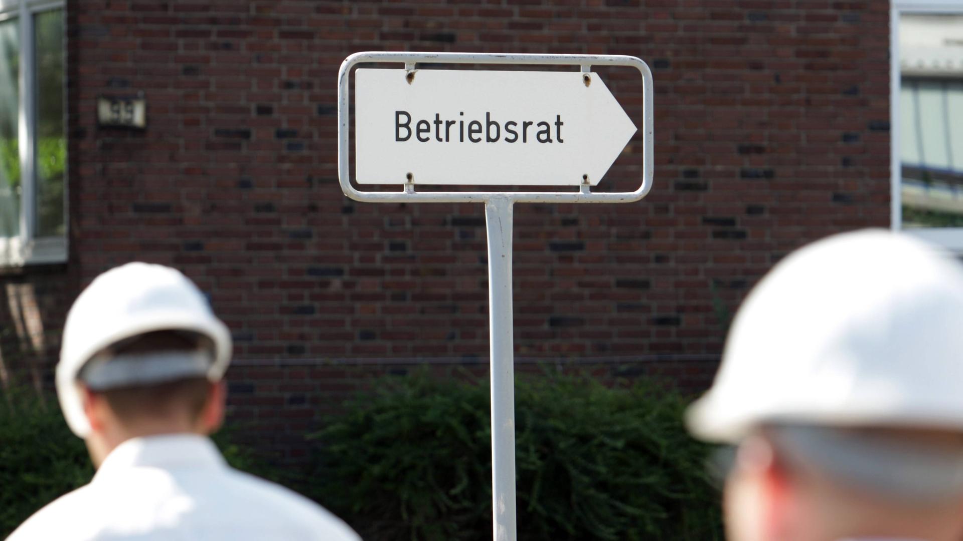 Arbeiter mit weißem Helm gehen an einem Schild vorbei, auf dem "Betriebsrat" steht.